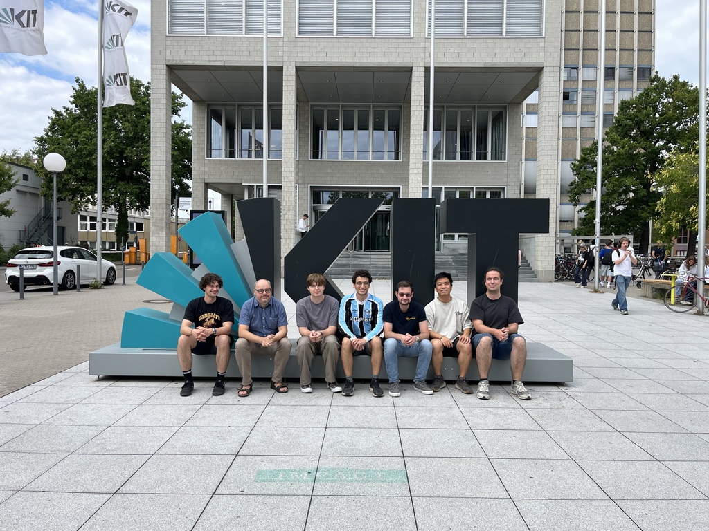 Six men sitting in front of a large KIT logo sculpture and university building.